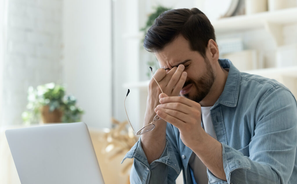 Tired young man feel pain eyestrain holding glasses rubbing dry irritated eyes fatigued from computer work, stressed man suffer from headache bad vision sight problem sit at home table using laptop.